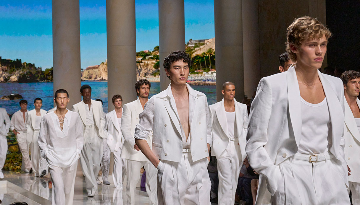 Modelli in giacche doppiopetto bianche aperte su t-shirt e pantaloni fluidi, sfilata Dolce&Gabbana Uomo SS27 con colonne e costa siciliana sullo sfondo