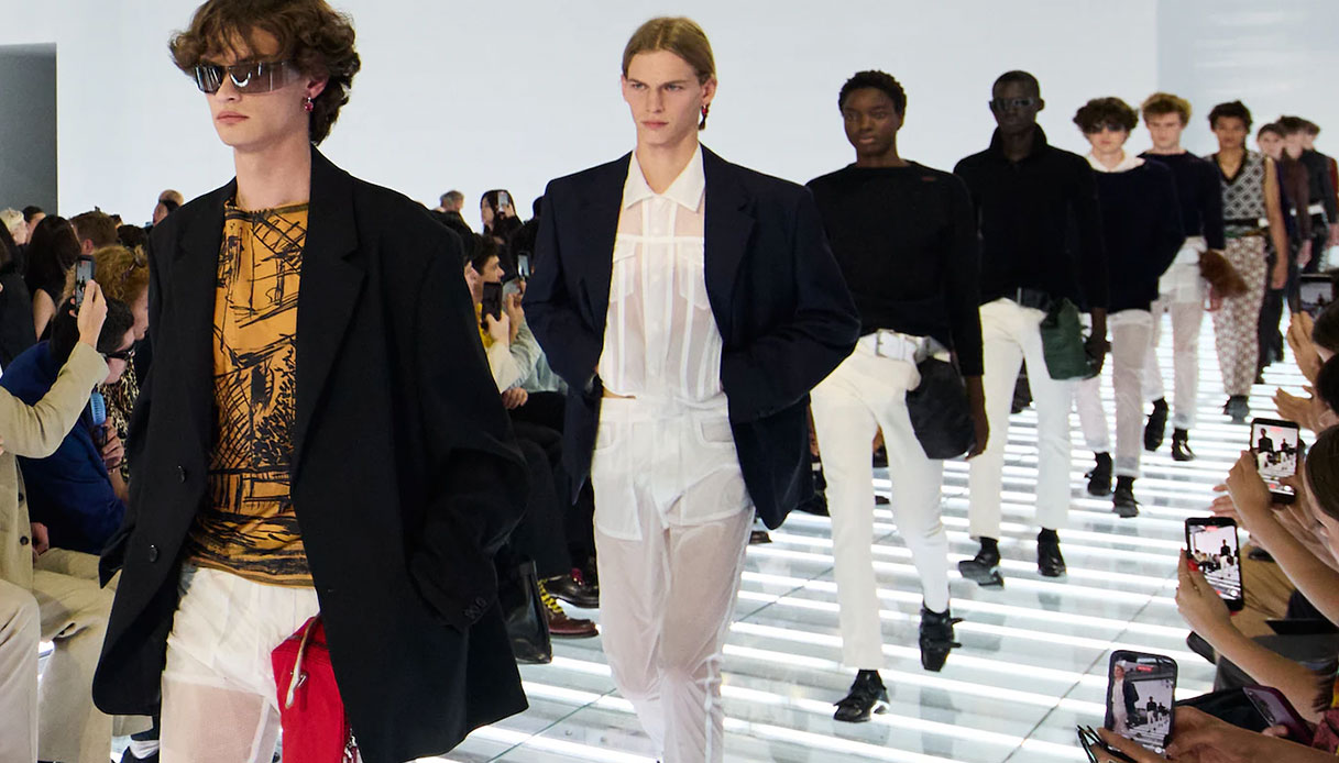 Modelli in blazer oversize nero su camicia trasparente e pantaloni bianchi sfilano sul palco luminoso del Deposito Fondazione Prada, SS27