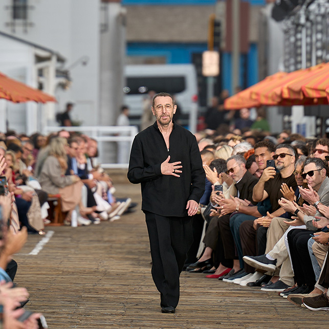 Alessandro Sartori in abito total black cammina sulla passerella del Malibu Pier tra il pubblico con ombrelloni arancioni sullo sfondo