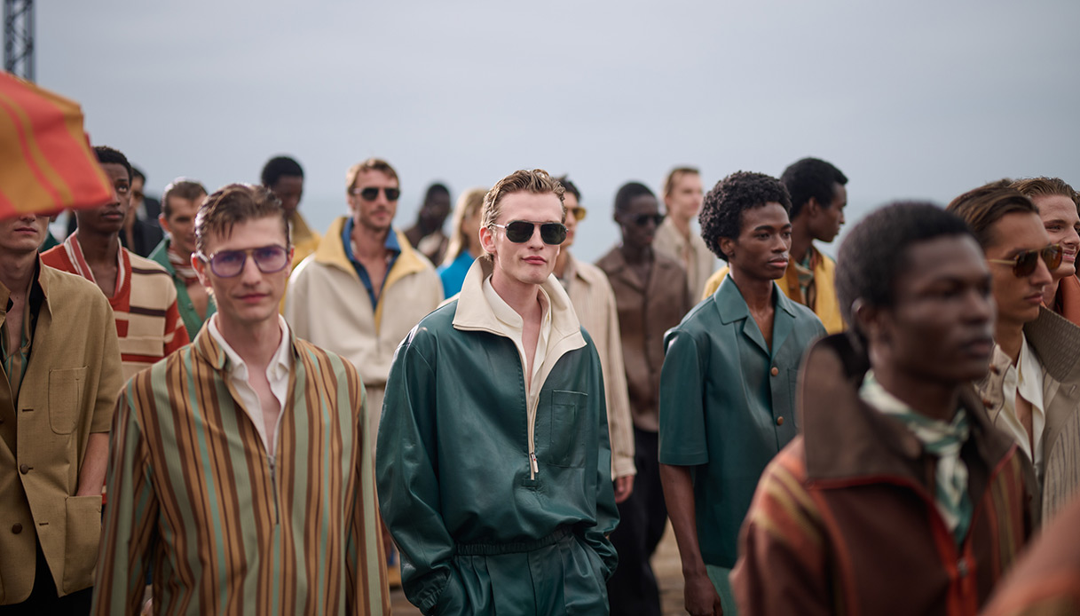 Gruppo di modelli sul Malibu Pier con look Zegna Estate 2027: tuta verde petrolio con colletto avorio in primo piano, attorno look in righe multicolore e toni sabbia