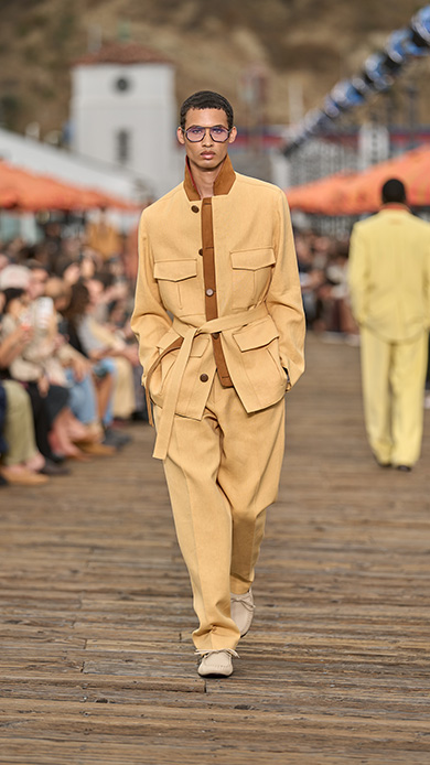 Modello con sahariana oversize color sabbia con cintura in vita, pantaloni ampi e scarpe basse crema sul Malibu Pier, ombrelloni arancioni sullo sfondo