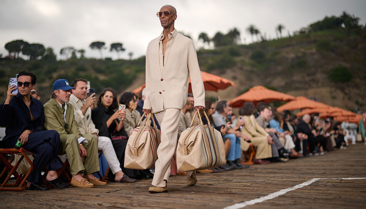 Modello in completo avorio con giacca a un bottone e pantaloni ampi porta due grandi borse in rafia a righe multicolore sul Malibu Pier