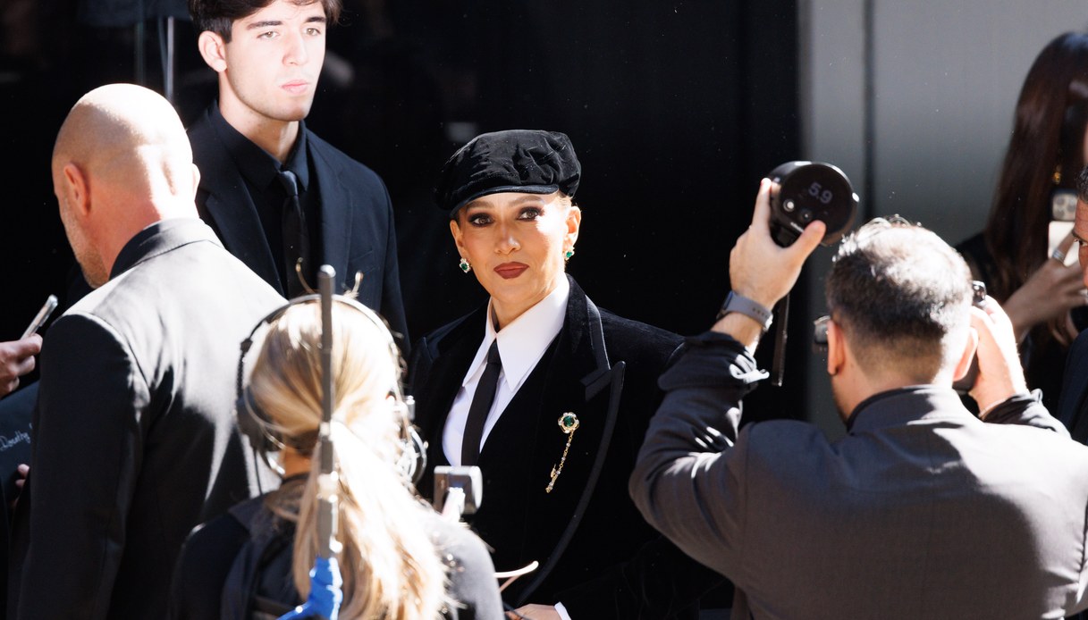 Jennifer Lopez in completo maschile di velluto nero con camicia bianca, cravatta, spilla gioiello e coppola, circondata da fotografi e operatori all'ingresso della sfilata