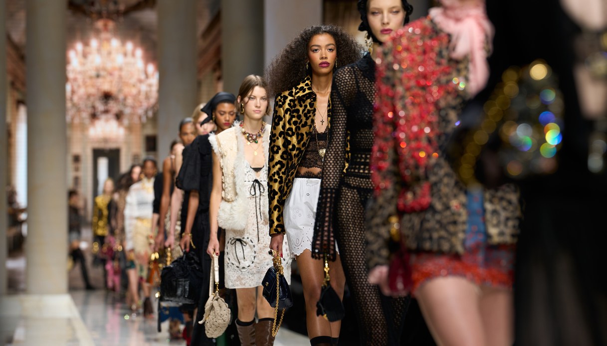 Fila di uscite in passerella con giacca animalier, abiti in pizzo e uncinetto, maglie a rete e borse gioiello, sotto i lampadari di una galleria con colonne