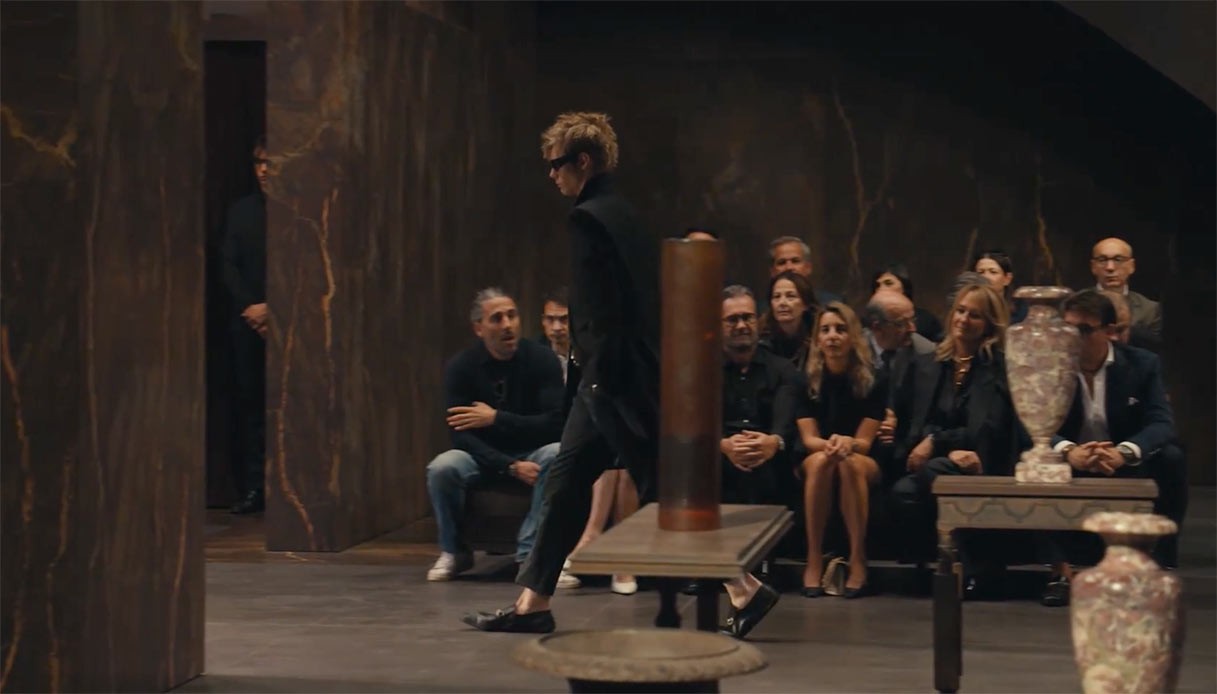 Uscita in total black che cammina in una sala rivestita in marmo scuro, tra vasi in ceramica su consolle e ospiti seduti lungo la parete