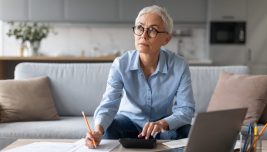 Donna con occhiali prende appunti su documenti, con una calcolatrice e un computer sul tavolo di casa.