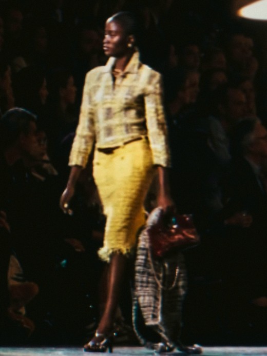 Rachel Ojuromi in passerella con camicia in tweed giallo e grigio, gonna midi gialla sfrangiata e borsa bordeaux in mano
