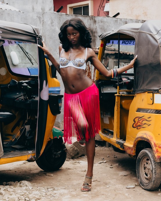 Rachel Ojuromi posa tra due risci&ograve; gialli in una strada di Lagos, con reggiseno ricamato, gonna a frange fucsia e sandali con tacco