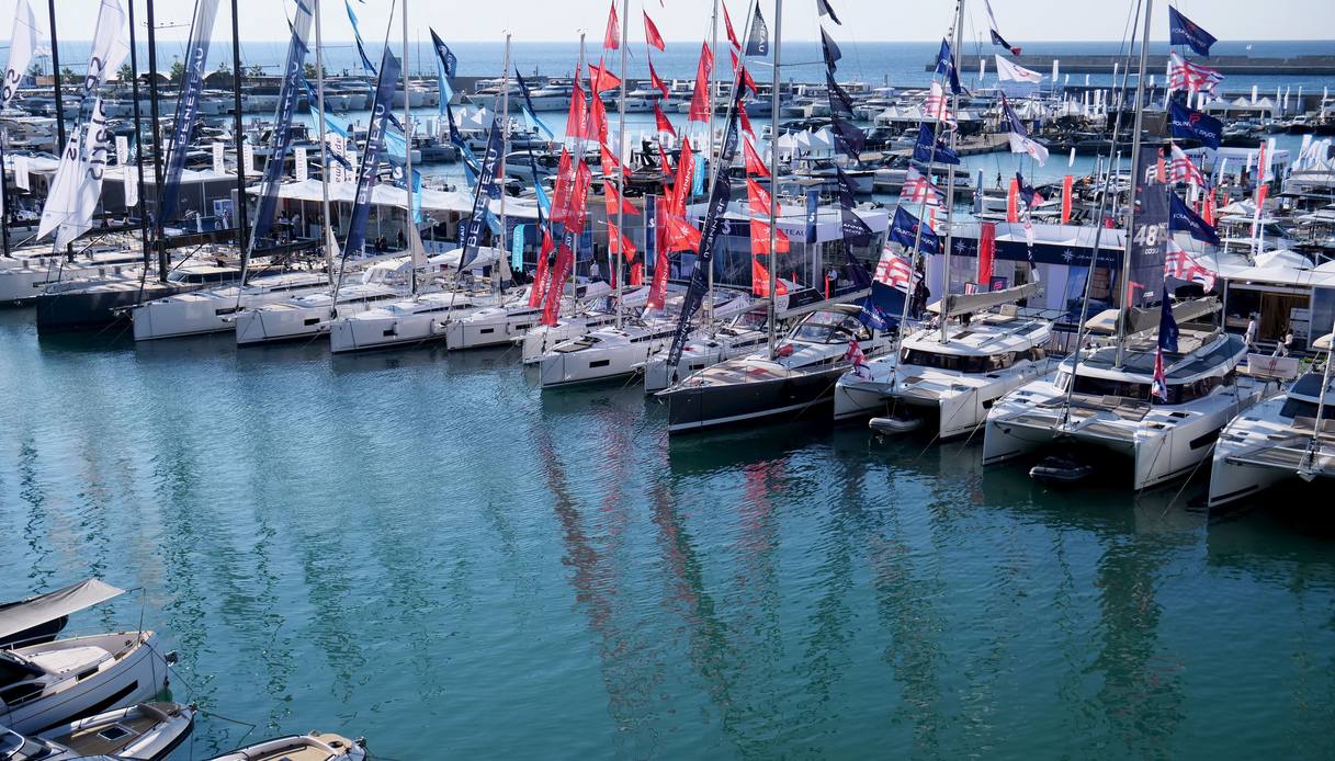 Veduta dall'alto delle banchine del Salone Nautico di Genova, con barche a vela e catamarani ormeggiati uno accanto all'altro, alberi e bandiere colorate degli espositori e il mare aperto sullo sfondo.